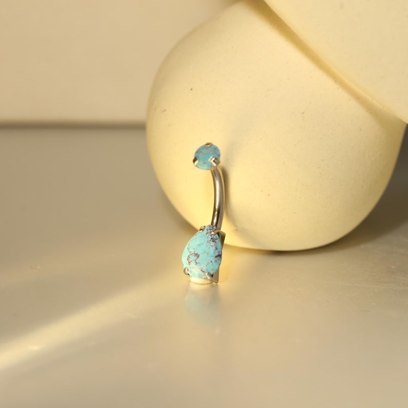 May include: A silver belly button ring with two turquoise-colored stones. The top stone is small and round, while the bottom stone is a larger, teardrop shape. The ring has a curved design.