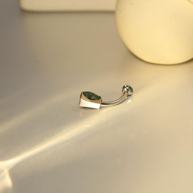 May include: A silver belly button ring with a curved barbell design. The ring features a square-shaped, green gemstone on the bottom and a small green gem on the top. The metal has a polished finish.