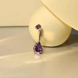 Puede incluir: Un piercing para el ombligo de plata con dos piedras preciosas moradas en forma de lágrima. La piedra más grande está en la parte inferior y la más pequeña en la superior. Las piedras son de color púrpura intenso y están engastadas en plata.