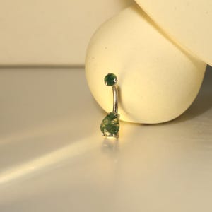 May include: A silver belly button ring with a curved barbell design. The top ball and the teardrop-shaped charm are both a deep green color. The charm has a textured, mossy appearance.