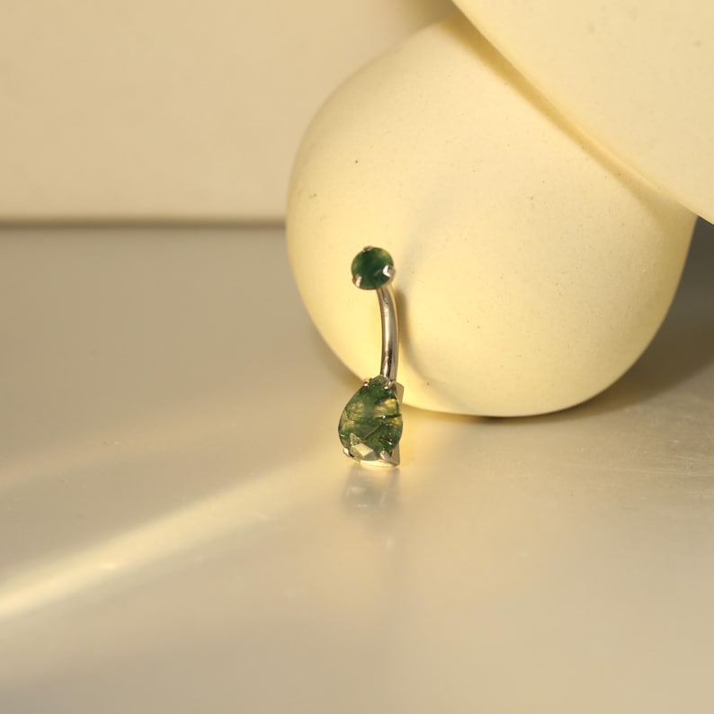 May include: A silver belly button ring with a curved barbell design. The top ball and the teardrop-shaped charm are both a deep green color. The charm has a textured, mossy appearance.