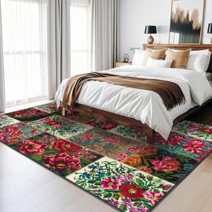 Può includere: Un tappeto patchwork colorato con motivi floreali in tonalità di rosso, rosa, verde e blu. Il tappeto è steso su un pavimento in legno chiaro in una camera da letto con un letto bianco e una coperta marrone.