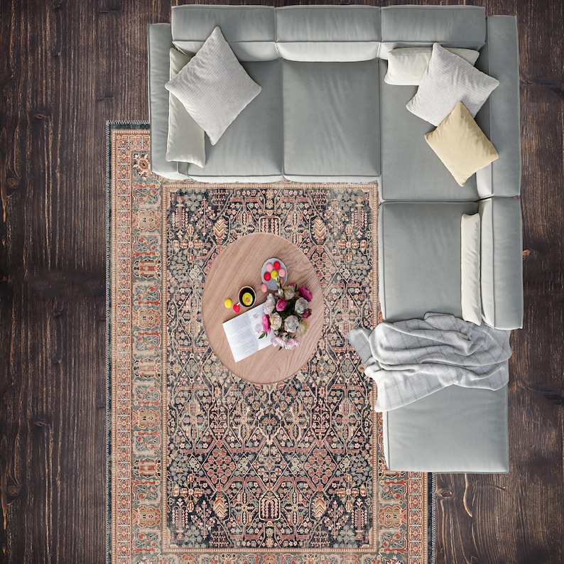 May include: Overhead view of a living room setup. A light gray sectional sofa with several throw pillows sits on a dark wood floor. A patterned rug and a round wooden coffee table with flowers and snacks complete the scene.