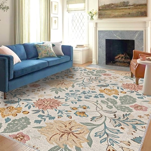 Puede incluir: Una alfombra con estampado floral sobre un fondo crema. La alfombra presenta grandes flores en tonos rosa, amarillo y azul, con hojas y enredaderas verdes. Un sofá azul y cojines son visibles en el fondo, creando un ambiente acogedor en el salón.