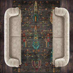 K&ouml;nnte beinhalten: Ein dunkelgrauer Teppich mit einem sich wiederholenden Muster aus stilisierten B&auml;umen in Blaut&ouml;nen, Gr&uuml;n, Rot und Gelb. Der Teppich liegt zwischen zwei beigen Sofas.