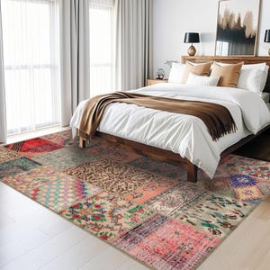 Op de afbeelding: Een patchwork tapijt met verschillende kleuren en patronen, waaronder rood, roze, blauw en groen. Het tapijt ligt in een slaapkamer met een wit bed en een bruin dekbed.