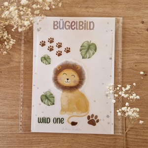 Puede incluir: Una transferencia termoadhesiva blanca con un lindo león de dibujos animados, hojas verdes y huellas de patas marrones. El texto "Wild One" está impreso debajo del león. El texto "Bügelbild" está impreso en la parte superior de la transferencia.