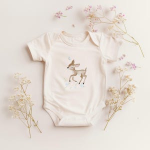 Könnte beinhalten: Cremefarbener Baby-Body mit einer Illustration eines niedlichen Rehs, einem Schmetterling und floralen Akzenten. Der kurzärmelige Body ist aus weichem Stoff gefertigt, ideal für Neugeborene und Babys. Zarte Blütenzweige umgeben den Body.