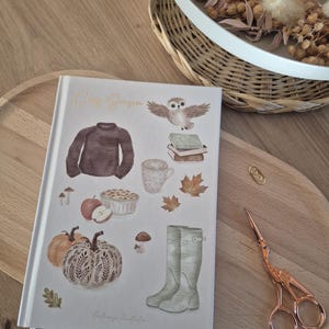 Könnte beinhalten: Ein weißes Notizbuch mit festem Einband, dem Text Cozy Season und Illustrationen eines braunen Pullovers, einer Eule, Büchern, einer Tasse, Herbstblättern, Pilzen, Äpfeln, einem Kuchen, Kürbissen und grünen Gummistiefeln sowie einem weißen Lesezeichen.