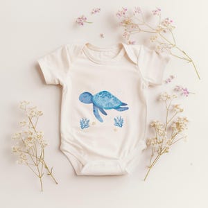 Op de afbeelding: Een witte babybody met een aquarel illustratie van een blauwe zeeschildpad. Het ontwerp bevat bubbels, koraal en zand. De bodysuit met korte mouwen is omgeven door witte bloemen met kleine roze en paarse bloesems.