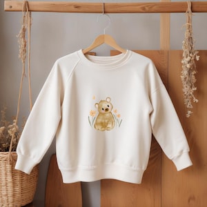 Könnte beinhalten: Cremefarbenes Sweatshirt mit einer Aquarellillustration eines braunen Bären, der zwischen gelben Blumen und grünen Blättern sitzt. Das langärmlige Oberteil hängt an einem Holzkleiderbügel und zeigt ein niedliches, skurriles Design für Kinder.
