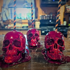May include: Three red skull-shaped candies with a glossy finish sit on a wooden surface. Each skull has a striped black and orange straw. The candies appear to be dripping. The image has a Halloween theme.