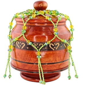Puede incluir: Un tarro de madera hecho a mano con tapa, adornado con un collar de cuentas. El tarro es de color marrón oscuro con un diseño decorativo de corazón. El collar de cuentas presenta cuentas amarillas y verdes, añadiendo color.
