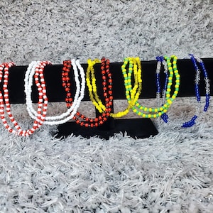 Puede incluir: Una exhibición de collares de cuentas de colores. Los collares están dispuestos sobre un soporte negro sobre un fondo gris texturizado. Los collares presentan cuentas rojas, blancas, amarillas, verdes, azules y transparentes. Cada collar parece estar hecho de múltiples hebras.