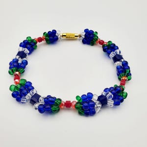 May include: A beaded bracelet featuring clusters of blue, green, and clear beads, interspersed with red beads. The bracelet has a gold-colored clasp and is set against a white background. The beads are arranged in a floral pattern.