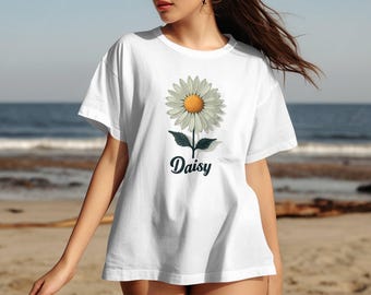 以下が含まれることがあります： 白いオーバーサイズのTシャツに、黄色い中心と緑の葉を持つ白いデイジーのグラフィックがプリントされています。花の下には「Daisy」という文字がプリントされています。
