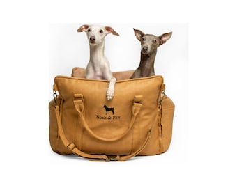 Può includere: Una borsa trasportino per animali color beige con due manici e una tracolla regolabile. La parte anteriore presenta una sagoma di cane ricamata in nero sopra la scritta Noah & Paw. Due levrieri italiani sono seduti all'interno.
