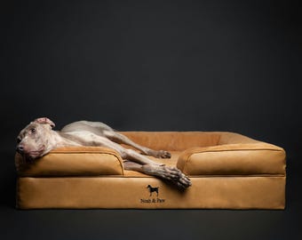 May include: A tan rectangular pet bed with raised sides and a smooth surface. A Weimaraner dog is lying on the bed. The front of the bed features a small black silhouette logo of a dog and the text Noah & Paw printed in black lettering.