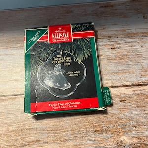 May include: A Hallmark Keepsake Ornament box from the Collector's Series. The box is green, red, and black, with text that reads "The Twelve Days of Christmas 1992." The ornament features nine ladies dancing. The box is on a wooden surface.