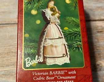 Könnte beinhalten: Eine rote und goldene Keepsake Ornament-Box mit einer viktorianischen Barbie-Puppe, die einen Teddybären hält. Die Box trägt den Text "Victorian BARBIE with Cedric Bear Ornament" und "DÉCORATION DES FÊTES". Der Hintergrund zeigt einen Weihnachtsbaum.