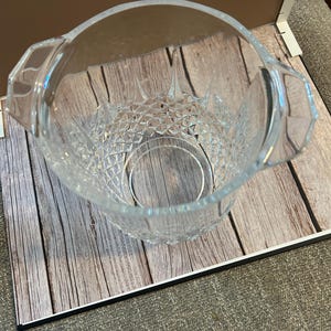 May include: A clear, faceted glass ice bucket with two handles. The bucket has a diamond pattern on the lower half and a smooth rim. The ice bucket is designed for serving beverages.