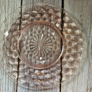 May include: A clear, pink-tinted glass plate with a diamond pattern and a raised center. The plate has a circular shape and is designed for serving food. The plate is made of glass and is likely vintage.