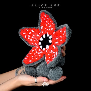 Peut inclure: Une peluche artisanale en forme de fleur de Demogorgon. La fleur a des pétales rouges avec des points blancs, un centre noir et des accents gris. Le jouet est posé sur un fond noir. Le texte "ALICE LEE HANDMADE" est visible en haut.