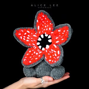 Pode incluir: Uma criatura Demogorgon de pelúcia feita à mão em vermelho, branco, preto e cinza. O Demogorgon tem uma grande cabeça em forma de flor com pétalas vermelhas salpicadas de pontos brancos. O texto "ALICE LEE HANDMADE" está no topo.