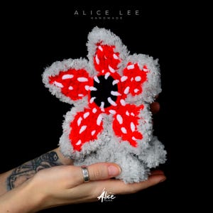 Pode incluir: Pelúcia artesanal de flor Demogorgon com pétalas vermelhas, pintas brancas e centro preto. A pelúcia é cinzenta e fofa, com o texto "ALICE LEE HANDMADE" no topo e "Alice" no canto inferior direito.