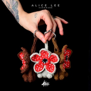 Pode incluir: Chaveiros de crochê feitos à mão com designs inspirados no Demogorgon. Os chaveiros são marrons, vermelhos e brancos, com formato de flor. A imagem inclui o texto "ALICE LEE HANDMADE" e a assinatura do artista.