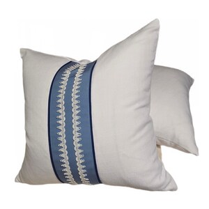 May include: Two white decorative throw pillows. Each pillow features a vertical blue ribbon with white lace trim down the center. The pillows are made of a soft fabric and are designed for home decor.