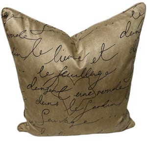 May include: A decorative pillow with a champagne-colored background and black script writing. The pillow has a soft, textured appearance and a classic design. The script appears to be French text. This pillow is a decorative home accent.