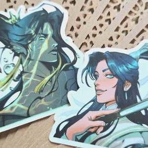 May include: Two stickers featuring anime-style illustrations of male characters with long dark hair and blue eyes. One character has a teal and gold color scheme, the other is in white and green. The stickers are on a decorative fan.