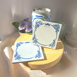 May include: Two square notepads with blue floral designs and the Japanese word "kawaii" are displayed on a wooden tray. A can of Sangaria soda and a small potted plant with purple flowers are in the background.