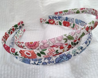 May include: Three floral headbands in shades of pink, red, and blue. Each headband is covered in a fabric with a different floral pattern. The headbands are arranged on a white, textured surface.
