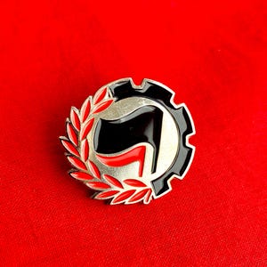 Peut inclure: Épinglette argentée avec un drapeau noir et un accent rouge. L'épinglette est entourée d'une bordure noire en forme de roue dentée et d'une couronne de laurier rouge et verte. Sur fond rouge.