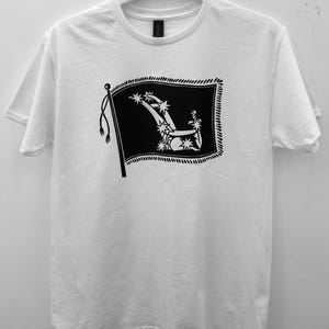 Könnte beinhalten: Ein weißes kurzärmeliges T-Shirt mit einem schwarzen Grafikdruck auf der Brust. Das Design zeigt eine rechteckige Flagge mit Fransenrand, auf der ein stilisierter Pflug und mehrere Sternformen abgebildet sind. Das T-Shirt hat einen Rundhalsausschnitt und hängt an einem Kleiderbügel.