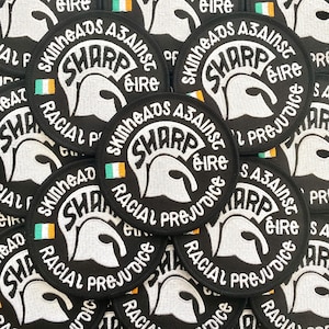 May include: A circular black embroidered patch featuring a white Trojan helmet graphic. The text reads SHARP, Skinheads Against Racial Prejudice, and Éire. A small green, white, and orange Irish flag is stitched on the left side of the helmet.