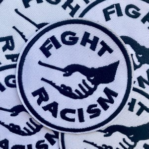 May include: A circular embroidered patch featuring a white background with a black border. The design displays a black silhouette of two hands shaking in the center, with the text FIGHT above and RACISM below in bold black capital letters.