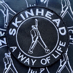 May include: A circular black embroidered patch featuring white stitching. The centre depicts a figure with one arm raised. The text SKINHEAD is arched across the top, and A WAY OF LIFE is arched across the bottom, all in white capital letters.