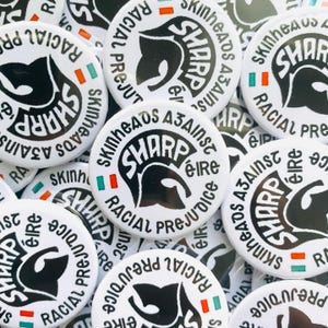Pode incluir: Uma coleção de botões brancos SHARP (Skinheads Against Racial Prejudice). Cada botão apresenta uma silhueta preta da cabeça de um skinhead, a palavra "SHARP" e o texto "Skinheads Against Racial Prejudice" em um design circular.