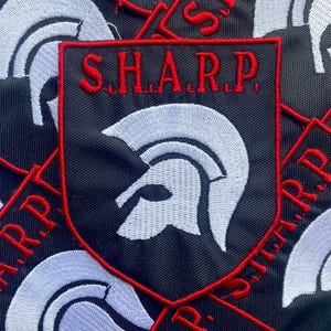 May include: A black shield-shaped embroidered patch featuring a white Spartan helmet icon in the center and the red text S.H.A.R.P. arched across the top. The patch has a red stitched border and is displayed against a background of identical patches.