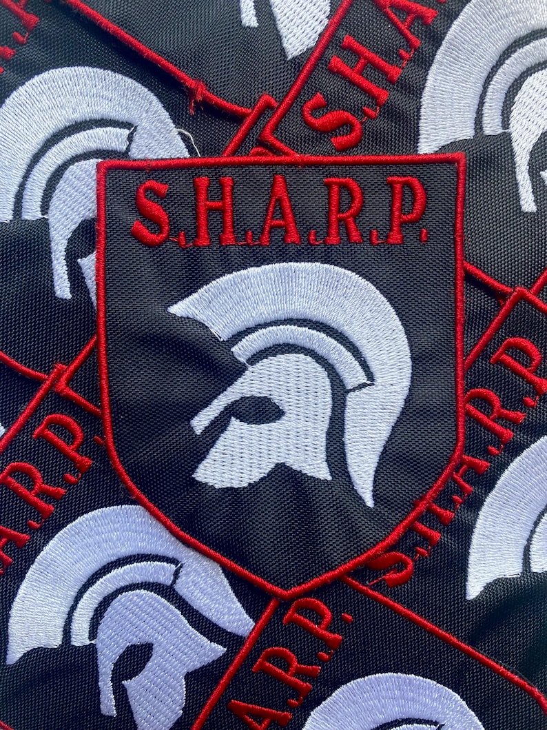 May include: A black shield-shaped embroidered patch featuring a white Spartan helmet icon in the center and the red text S.H.A.R.P. arched across the top. The patch has a red stitched border and is displayed against a background of identical patches.