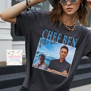 May include: Dark gray t-shirt with a graphic of two men and the text "CHEF BEN Below Deck" in blue. The image on the shirt shows a yacht in the background. The shirt is worn by a person wearing a white hat and sunglasses.