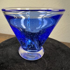 May include: A vibrant blue glass bowl with a wide, flared top and a rounded base. The bowl's interior is a deep blue, with visible air bubbles, and the rim is clear. The design is reminiscent of a cocktail glass.