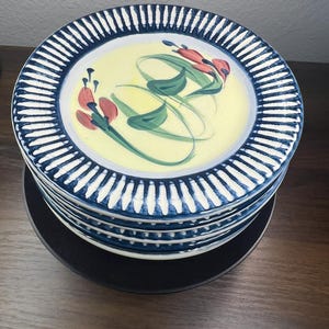 May include: A stack of four round ceramic plates featuring a pale yellow center decorated with hand-painted green leaves and red flower buds. The wide rims are adorned with a dark blue and white striped pattern, creating a bold contrast.