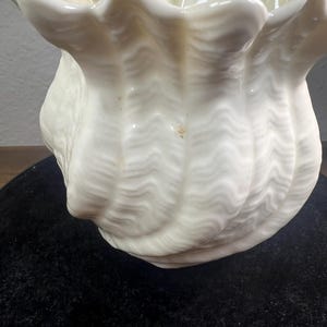 May include: A white ceramic seashell-shaped decorative bowl. The bowl has a ruffled edge and a textured, wavy surface. The interior has a light yellow hue. The bowl is sitting on a black surface.