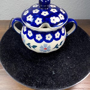 May include: A ceramic sugar bowl with a lid, featuring a blue and white floral pattern. The bowl has two handles and a small opening for a spoon. The lid is topped with a round knob. The bowl is approximately 4 inches in diameter.