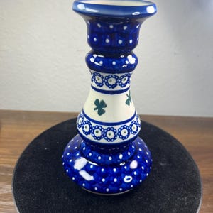 May include: A blue and white ceramic candlestick holder with a bulbous base and a flared top. The candlestick features a pattern of white dots and green shamrocks. The base is approximately 3 inches in diameter.