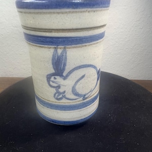 May include: A cylindrical ceramic tumbler with a speckled off-white base, featuring horizontal blue bands around the top and bottom. A hand-painted blue rabbit illustration is centered on the exterior surface of the vessel.